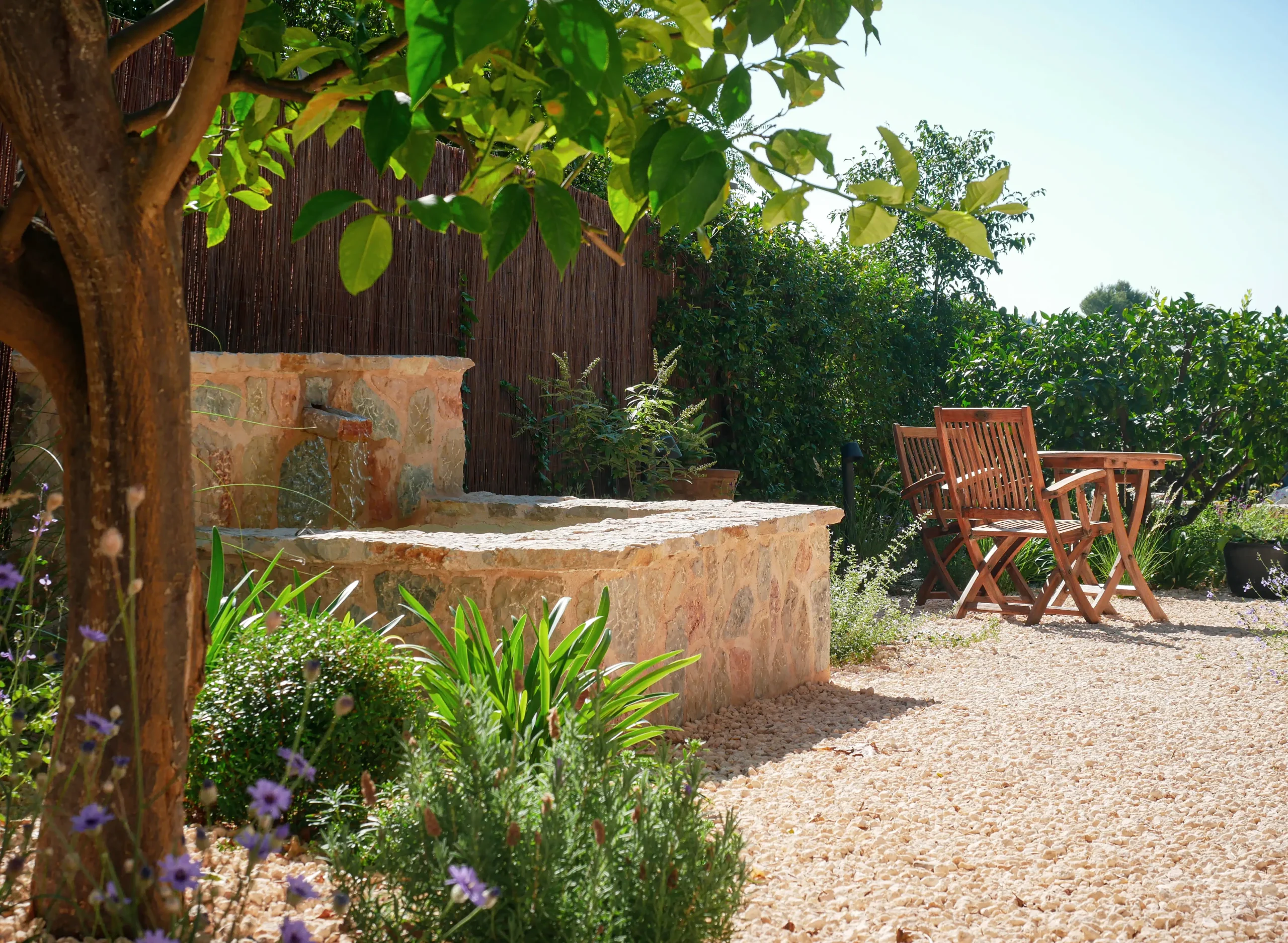 Sunlit patio with a rustic stone fountain, gravel ground, and Mediterranean planting.