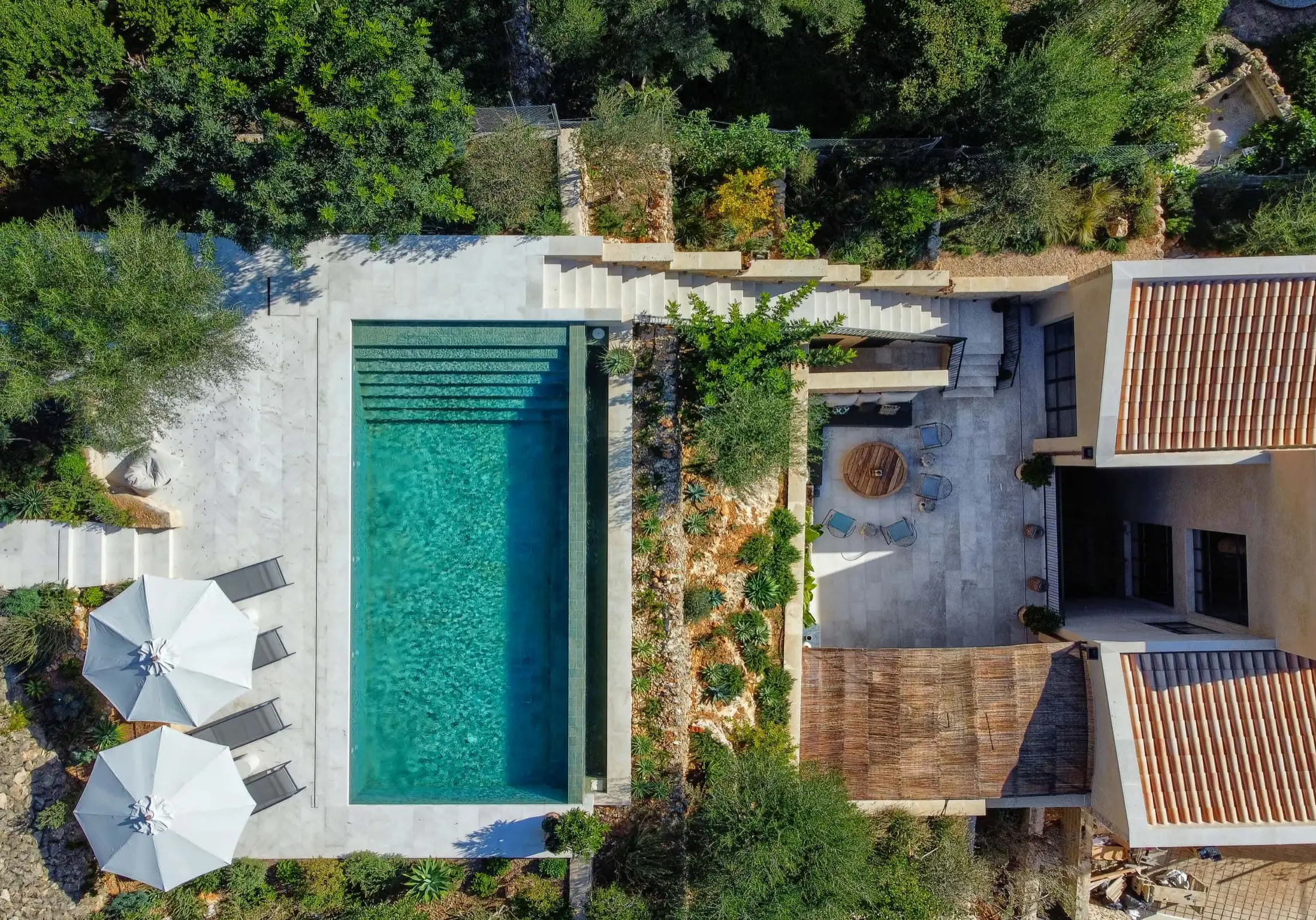Aerial view of a Mallorcan villa with stone terrace, rectangular pool, parasols, and surrounding Mediterranean garden