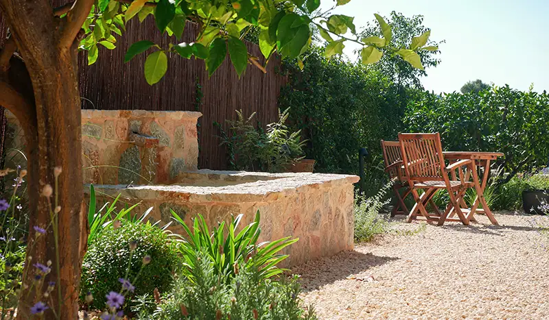 Sunny patio with a rustic stone fountain, wooden chairs, and Mediterranean-style garden plants