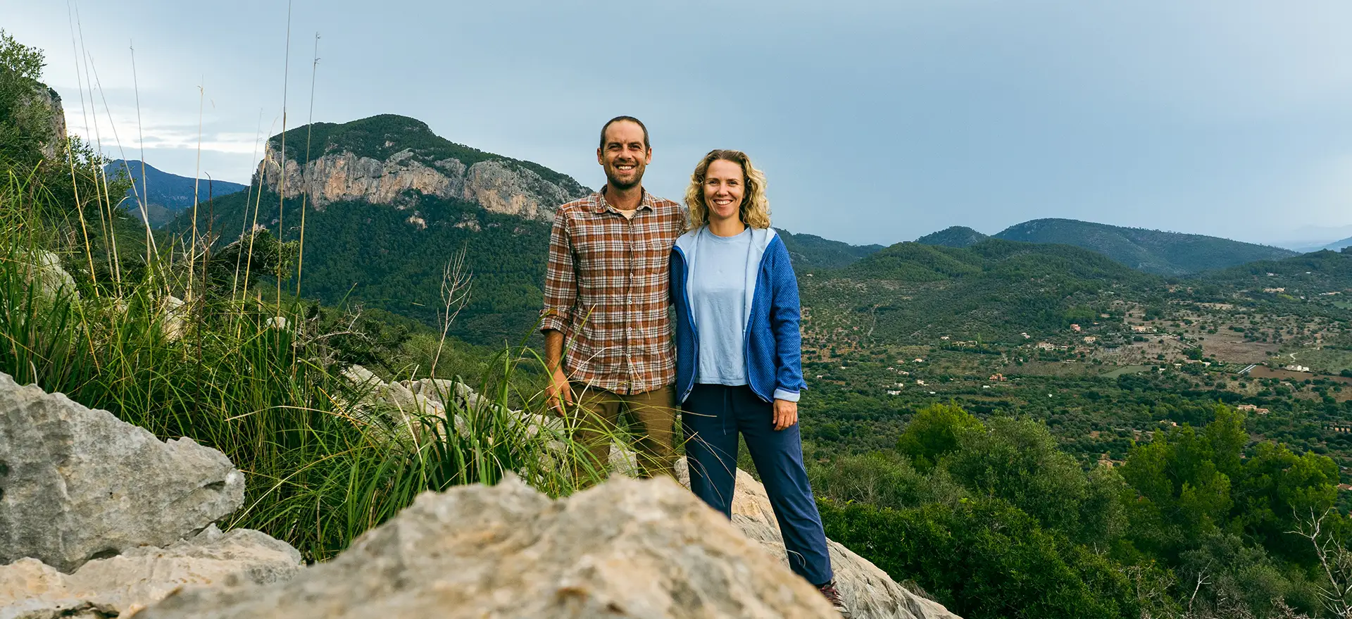 Jack and Jess on a rocky hillside with lush green valley and mountains behind them.